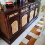  Un grand buffet en bois foncé de style classique, doté de quatre portes et de trois tiroirs, est présenté dans un intérieur. Les façades des portes et des tiroirs sont ornées de placages clairs à motifs marbrés. Le plateau du buffet est également en bois foncé et supporte divers objets décoratifs.