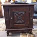 Une table de chevet ancienne en bois sombre, richement sculptée, est placée sur un tapis à motifs orientaux. Le meuble présente une porte centrale ornée d'un médaillon circulaire sculpté et encadré de motifs géométriques, flanqué de part et d'autre de panneaux verticaux également sculptés avec des motifs floraux circulaires