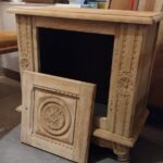 Une table de chevet ou petit meuble en bois clair, probablement après un décapage, est présentée de trois quarts face. La porte frontale sculptée, ornée d'un médaillon central et de motifs floraux sur les montants, est détachée ou ouverte et posée devant l'ouverture sombre du meuble. Le bois brut met en évidence les détails des sculptures. 