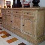  Un grand buffet en bois clair, probablement décapé ou restauré pour révéler sa couleur naturelle, est présenté dans un intérieur. Il comporte quatre portes et trois tiroirs supérieurs. Les façades des portes et des tiroirs sont ornées de placages clairs à motifs mouchetés, contrastant légèrement avec le reste du meuble.