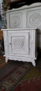Ensemble de chambre à coucher de style provençal ou shabby chic, comprenant une tête de lit et une table de chevet assortie, tous deux patinés dans des tons de blanc cassé. Les meubles sont ornés de motifs sculptés circulaires et floraux en relief, soulignant l'aspect vieilli et élégant.